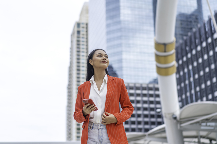 Confident Asian Businesswoman Holding Phone in City. Young Professional Female in Red Blazer Looking Ahead with Ambition