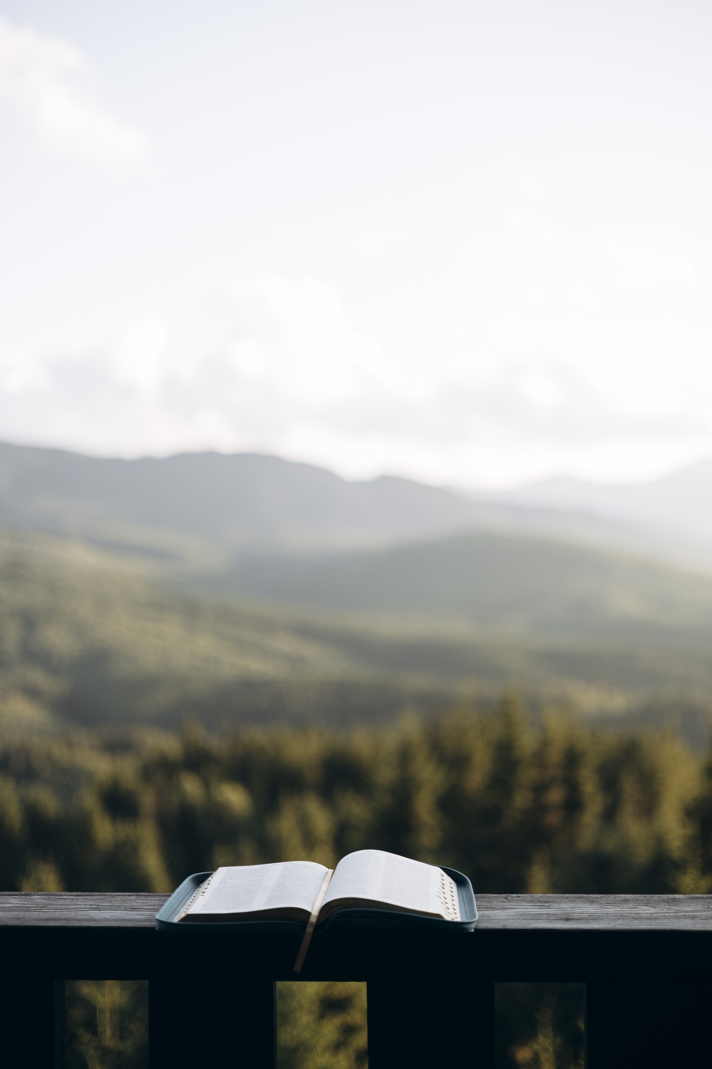 Open Bible on wooden balcony with mountain view and soft natural light.