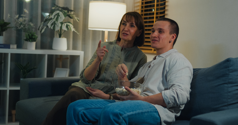 Caucasian mother and son relaxing together on the sofa while watching TV and eating popcorn in living room in home at night. Family lifestyle concept.